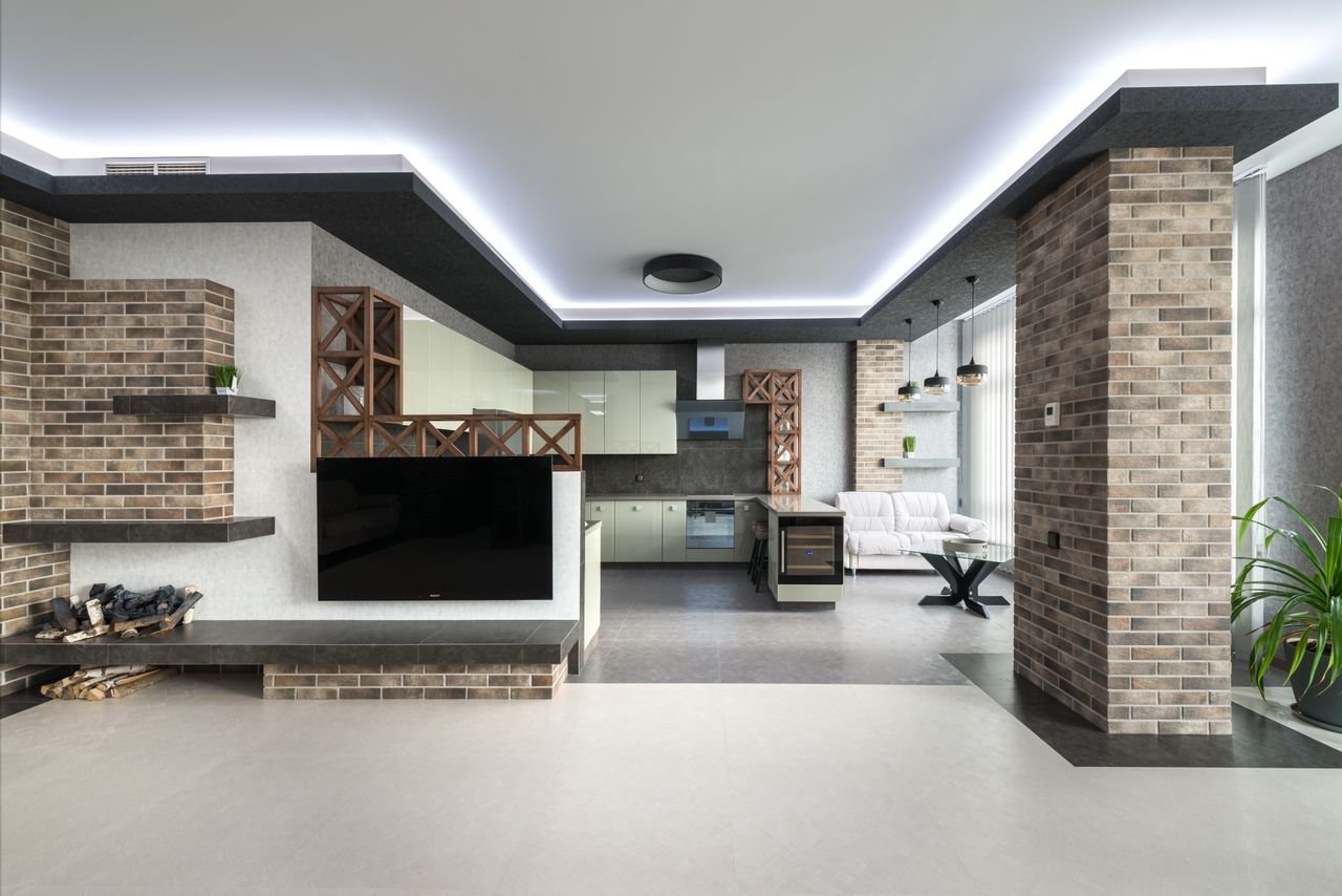 Kitchen with white cupboards placed near couch and modern TV in stylish apartment with pile of firewood and decorative brick column